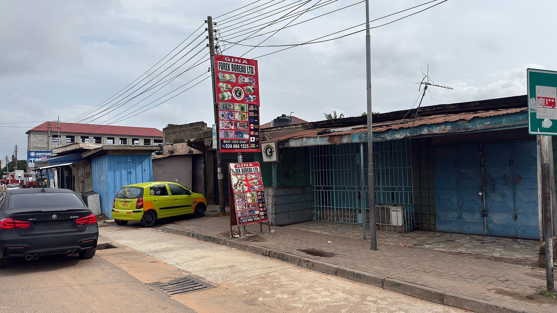A forex bureau sign displaying foreign currency notes beside a Ghanaian street with a taxi parked outside